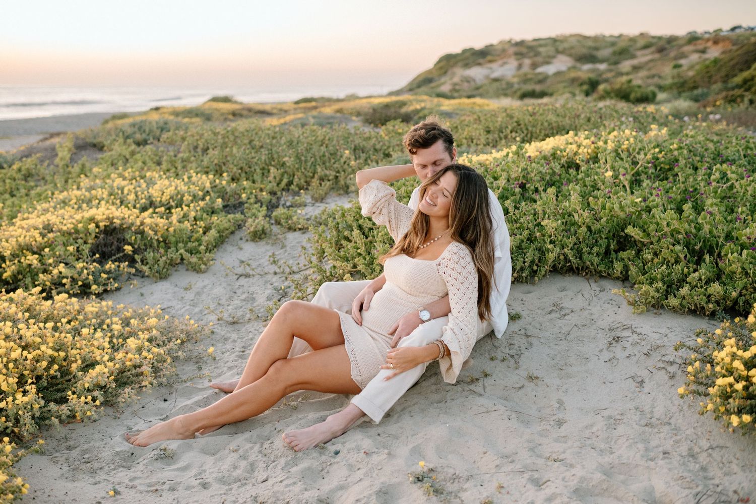 Couple relaxing together on sandy dunes surrounded by yellow wildflowers at sunset.