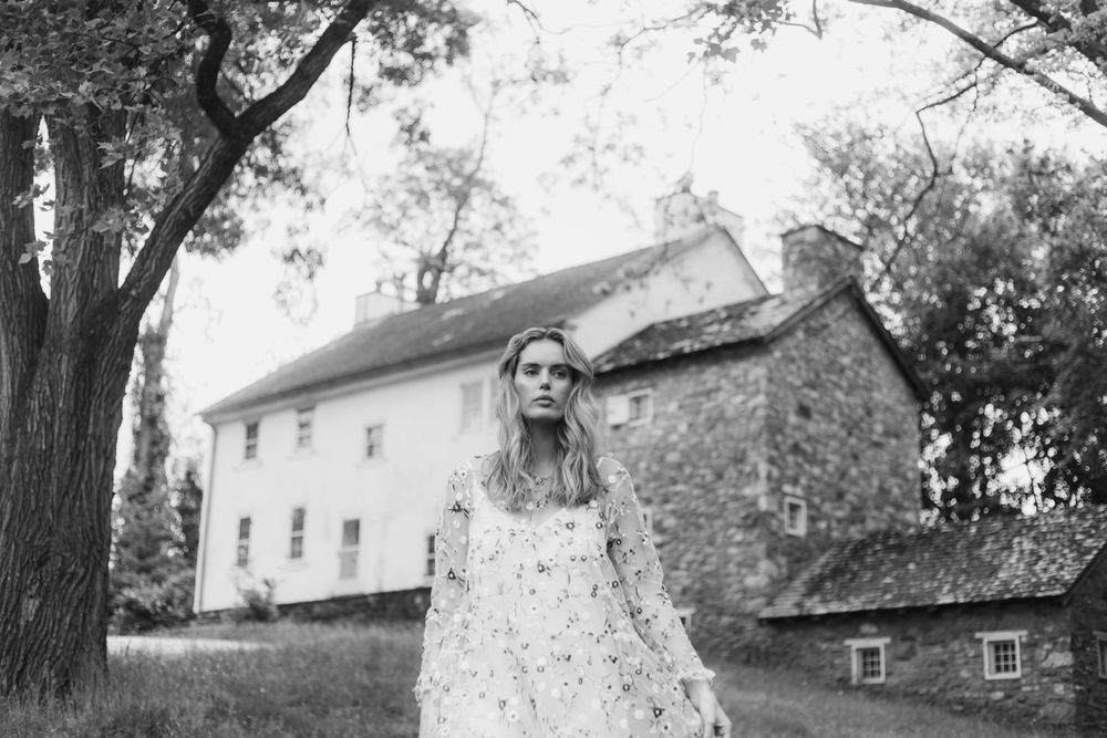 A vintage black and white photograph shows someone in a lace dress standing near a rustic stone farmhouse.