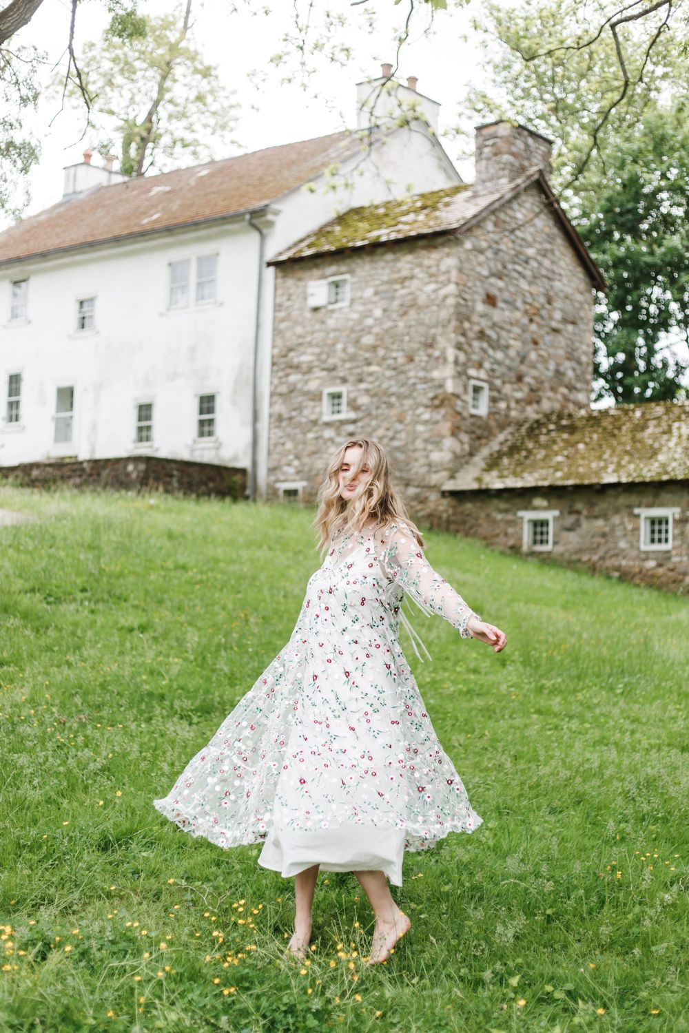 A light floral dress twirls on a grassy lawn in front of a historic stone and white farmhouse on a spring day.