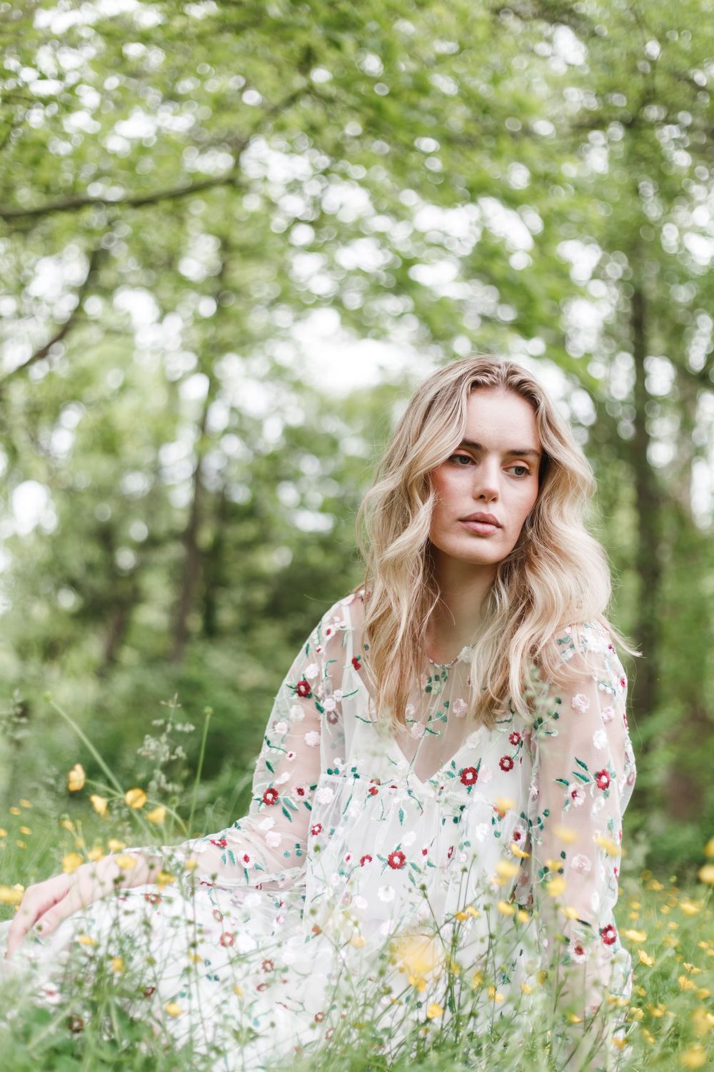 A woman in a floral white dress sits in a meadow of wildflowers with soft bokeh and trees in the background.