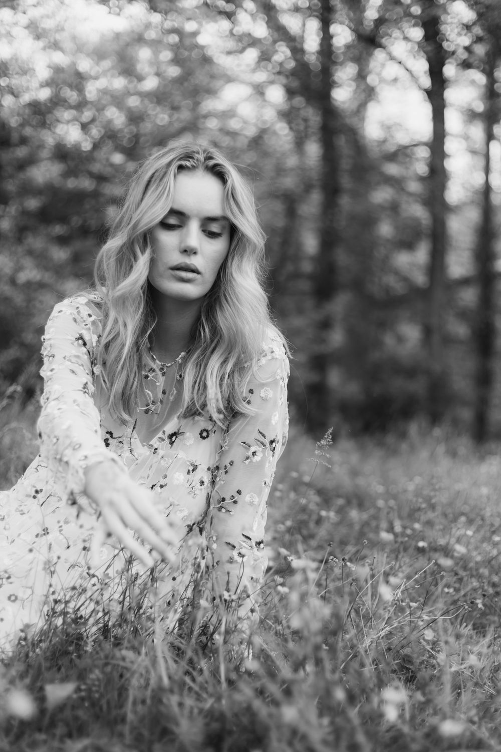 Black and white portrait of a woman wearing a delicate floral dress sitting in a dreamy meadow setting with trees in background.