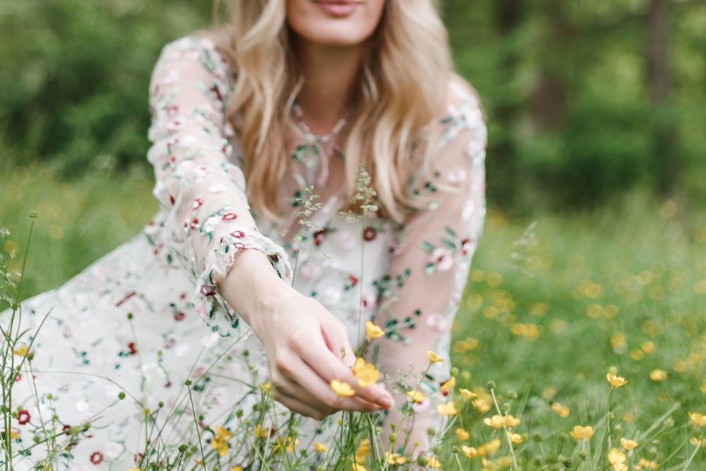 Woman in delicate lace floral dress picks wildflowers in a sunny spring meadow.