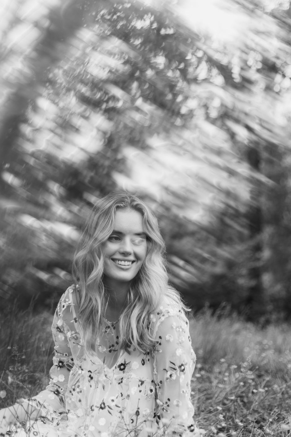 Black and white portrait of a person in a floral dress sitting outdoors in a natural, dreamy setting.
