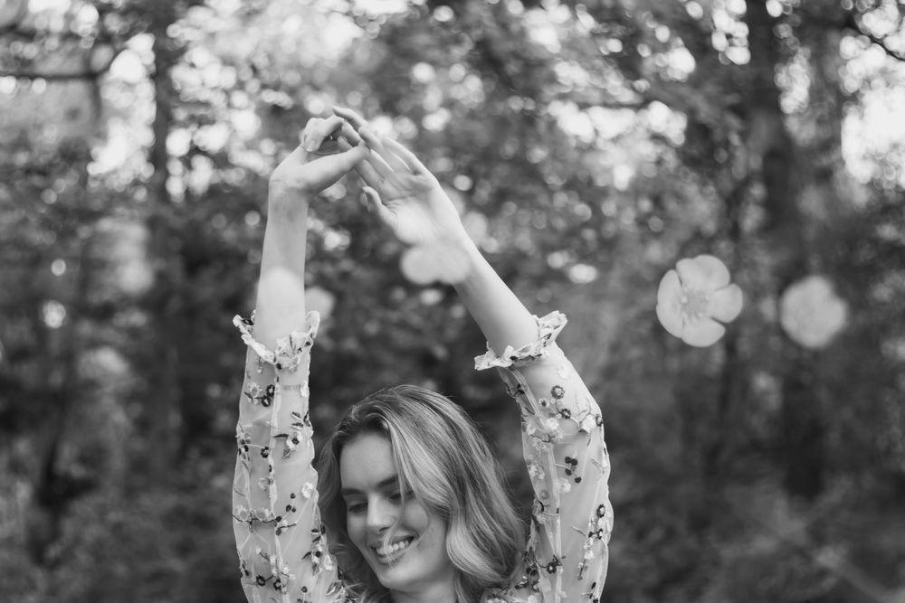 Black and white portrait of woman in floral dress dancing with raised arms in forest setting.