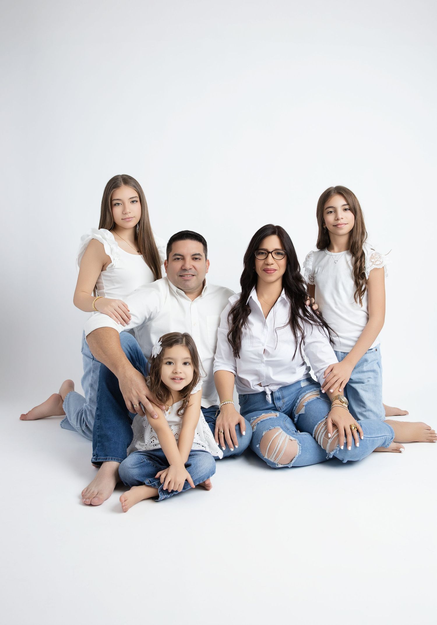 A family wearing blue jeans and white tops sits together barefoot against a plain white background.