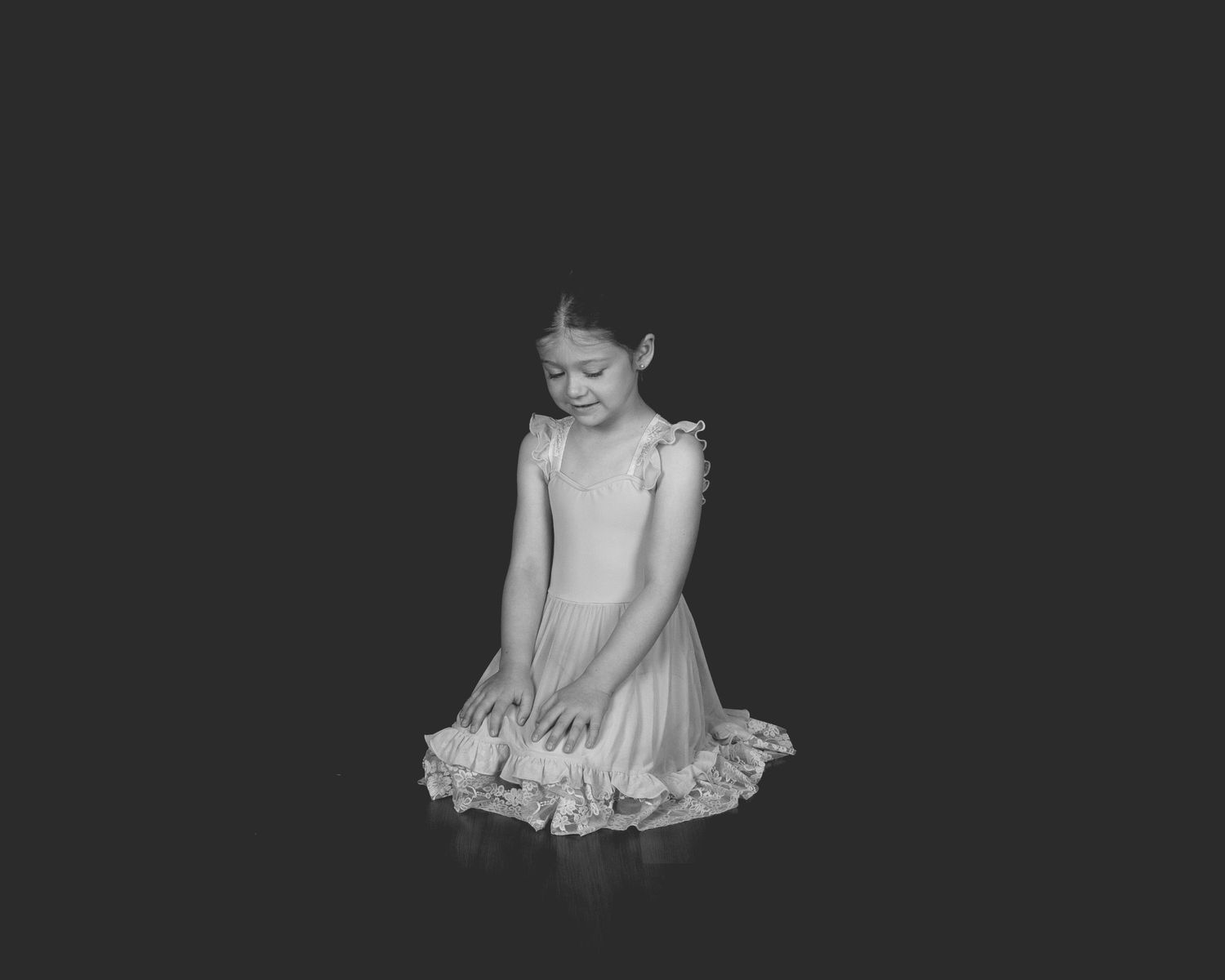 A young ballet dancer in a light-colored dress stands gracefully looking down in a black and white portrait.
