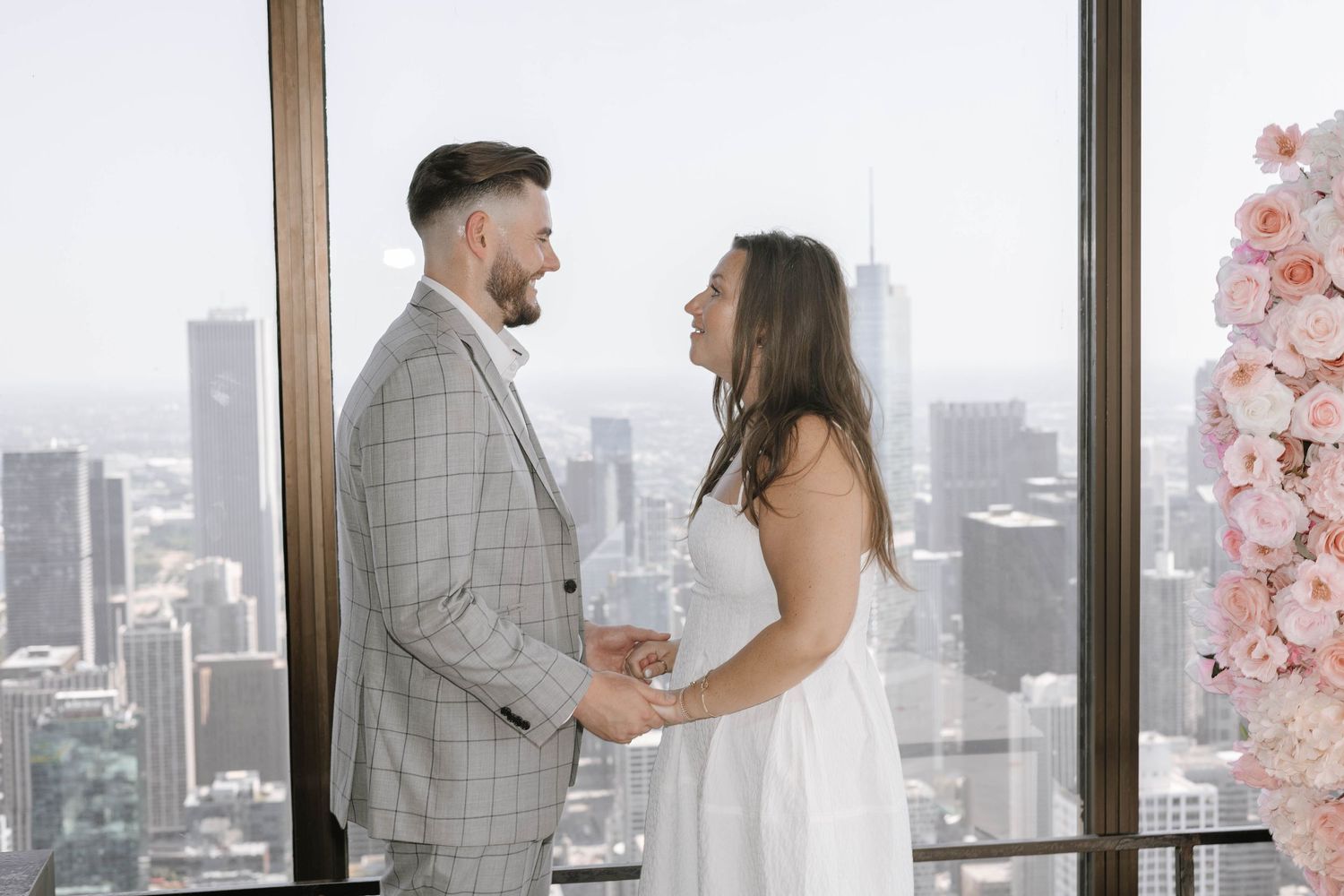A couple shares an intimate moment in front of floor-to-ceiling windows overlooking a city skyline with pink floral decor.