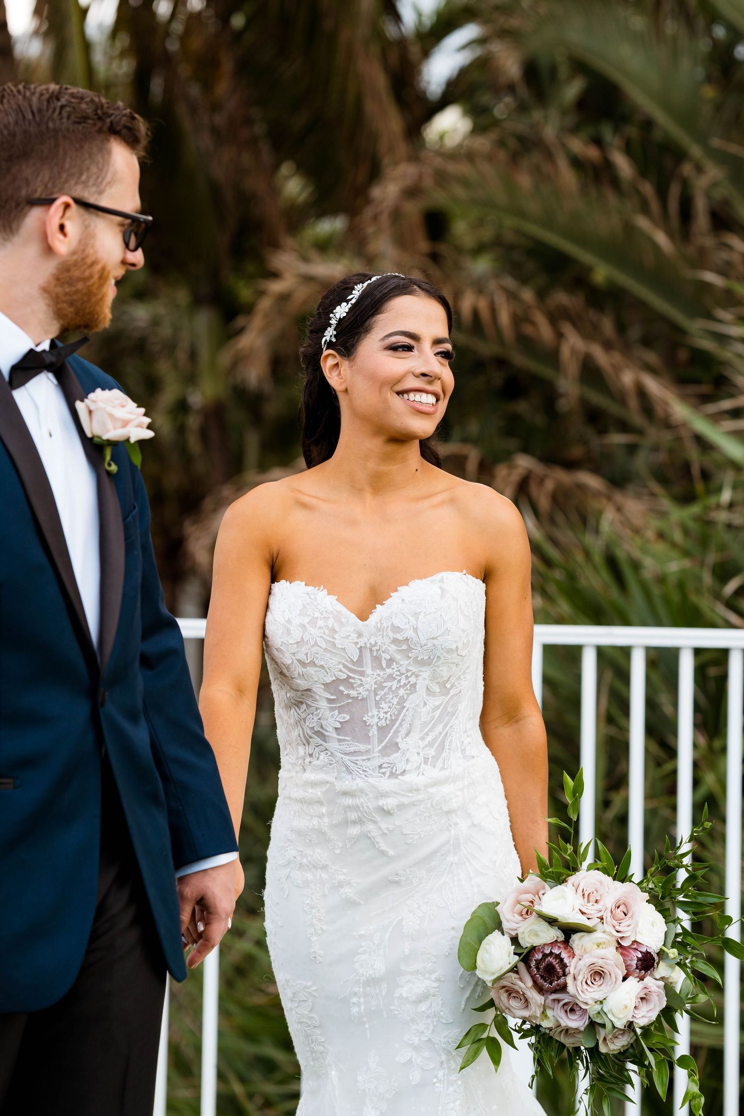 A wedding couple poses outdoors with tropical foliage and a beautiful bridal bouquet.