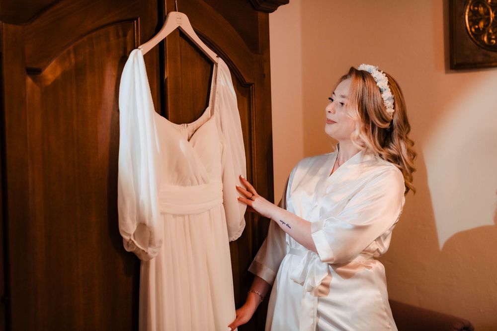 A white wedding dress hangs on a wooden wardrobe while someone in a silk robe admires it.