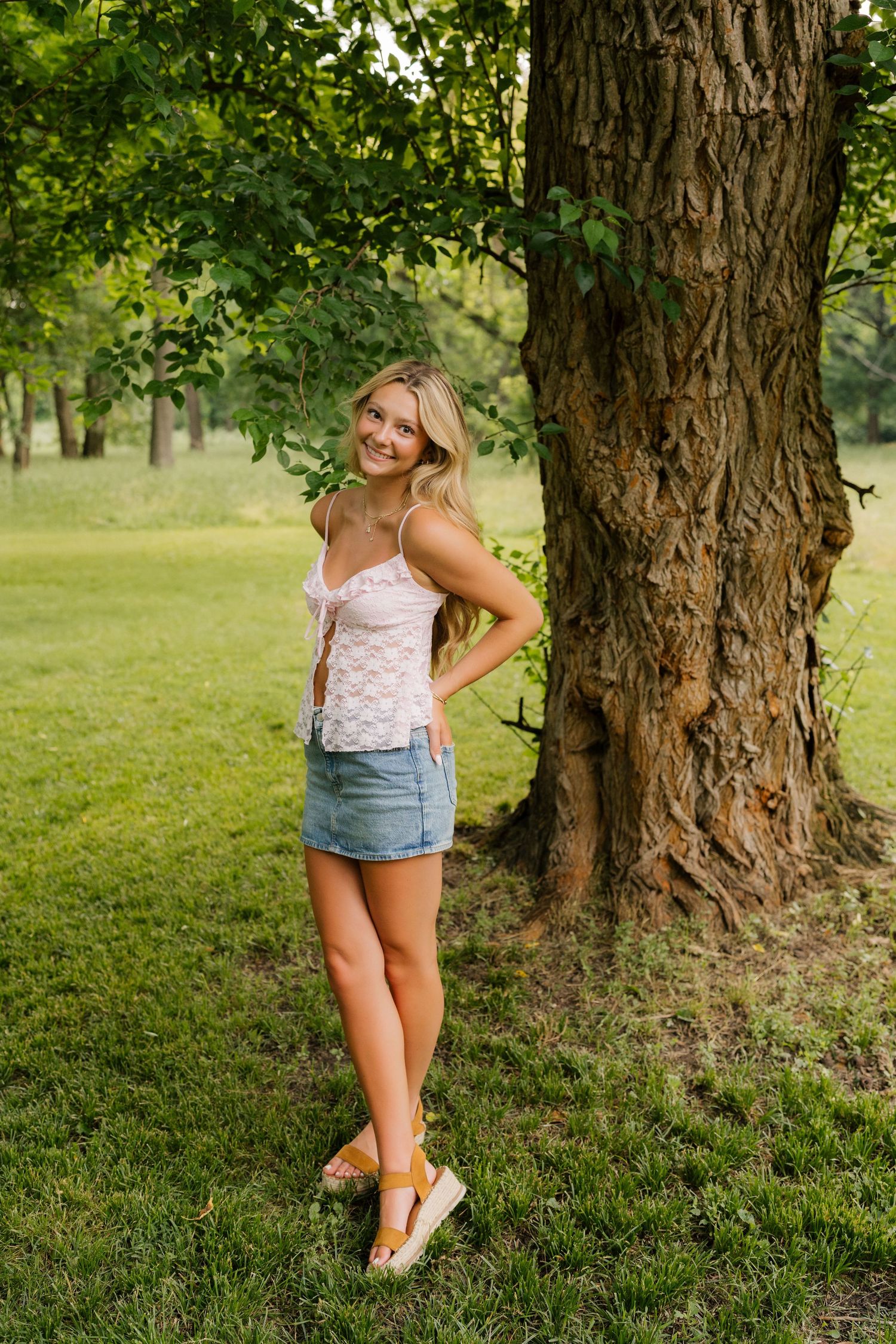 Figure leaning against large tree trunk in grassy park area wearing casual summer outfit on sunny day.