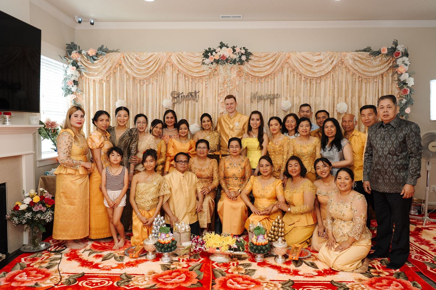 A large group wearing traditional gold and beige formal attire poses together at an elegantly decorated celebration event.