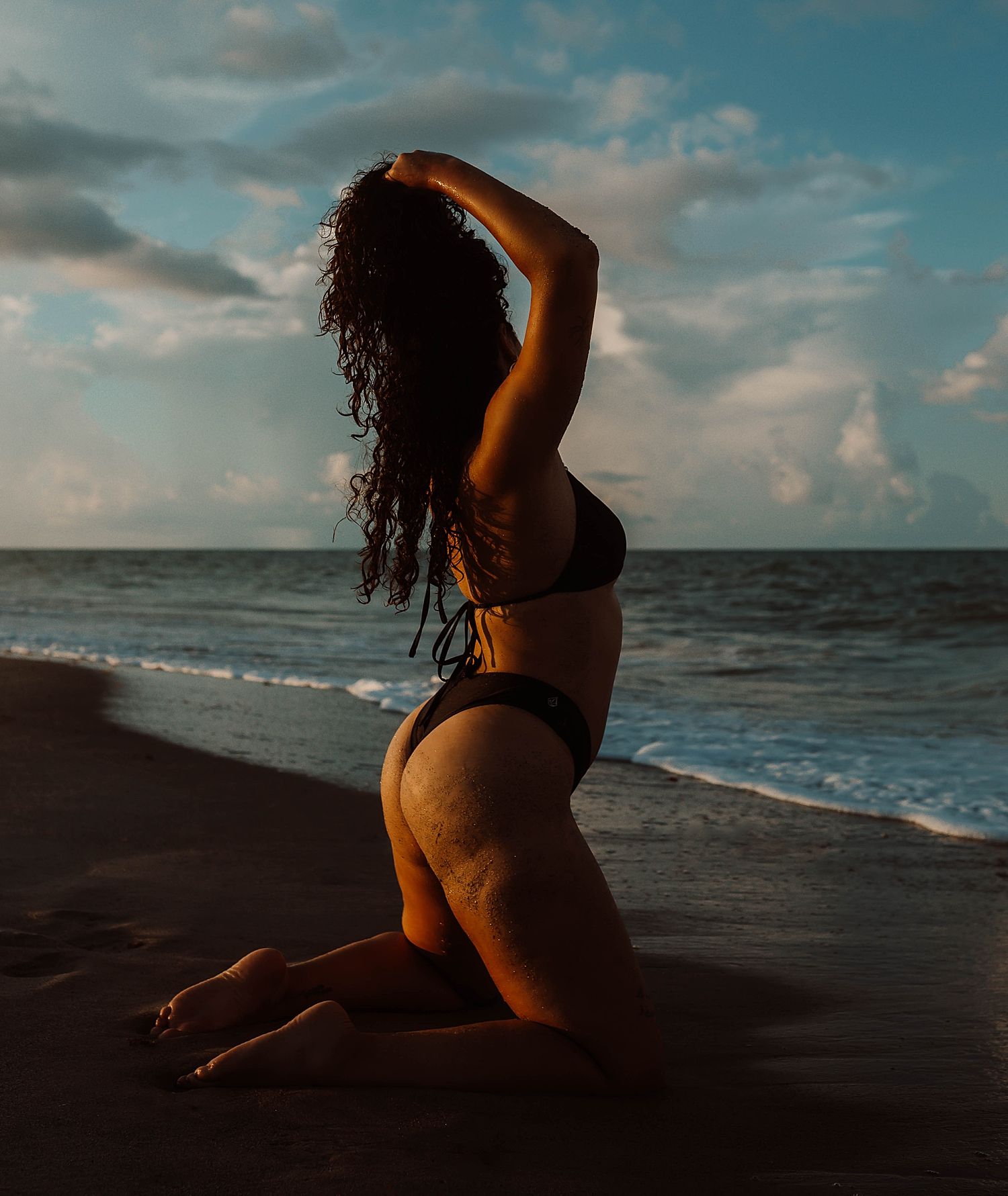 Woman in swimsuit poses on dark sandy beach at sunset by Wisconsin boudoir photographer