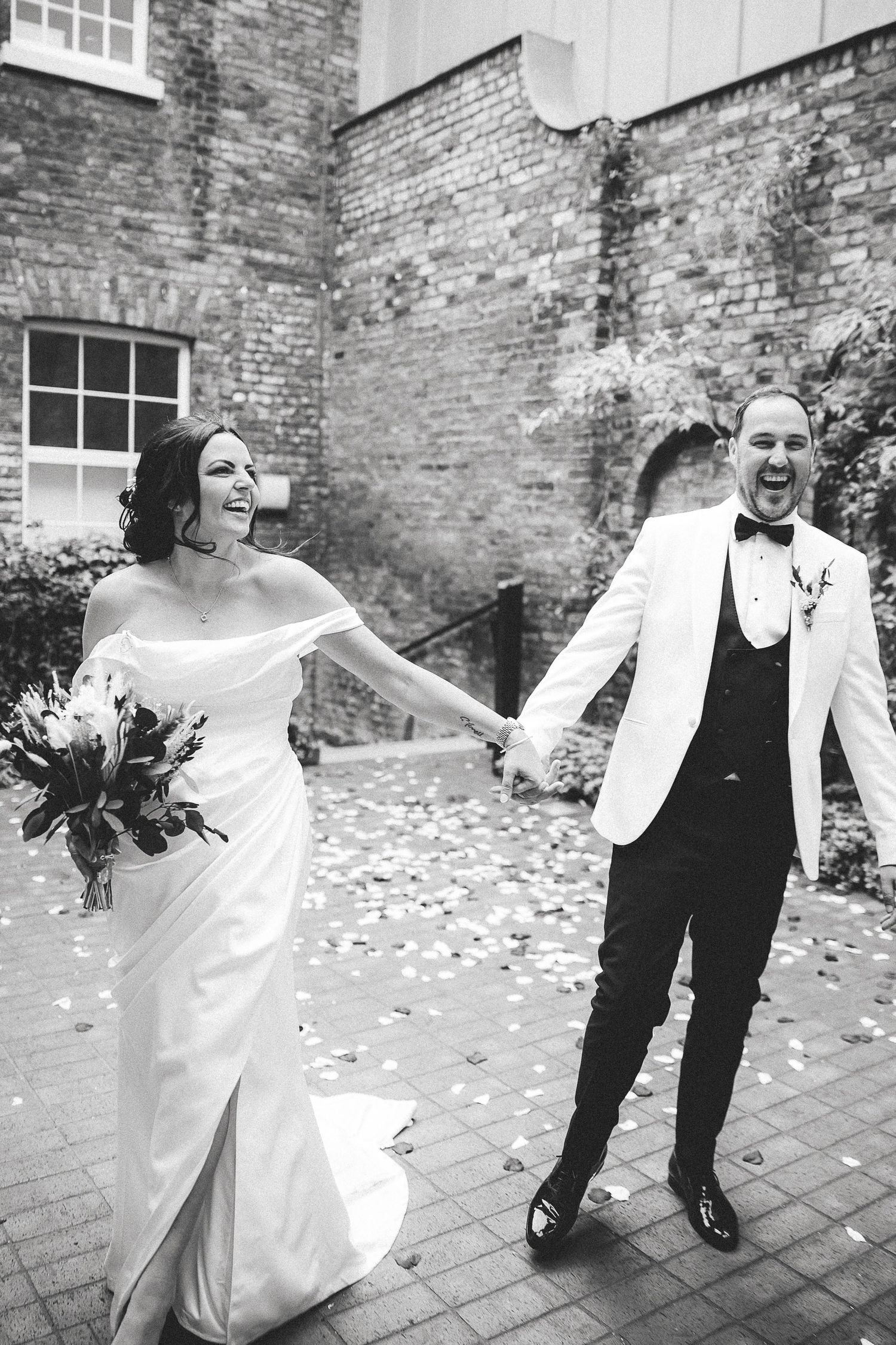 A joyful wedding couple holds hands and laughs while rose petals fall around them on a brick courtyard.