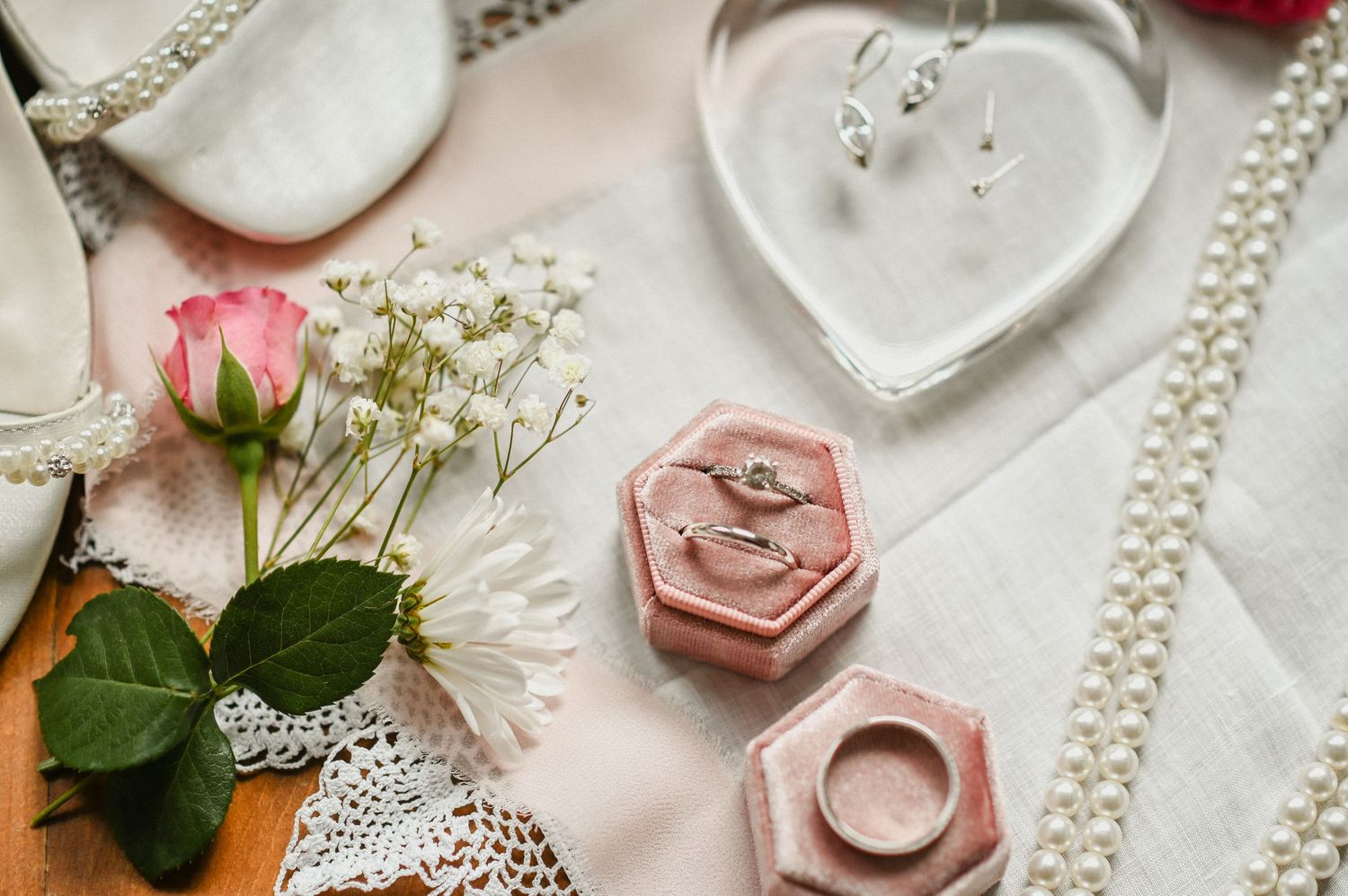 An image of a wedding vow book surrounded by a bouquet, perfume, eucalyptus leaves, ribbon, a ring box with rings, and shoes for a wedding.