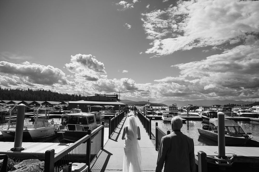 A black and white scenic view of a couple walking along a marina boardwalk with boats and clouds in the background.