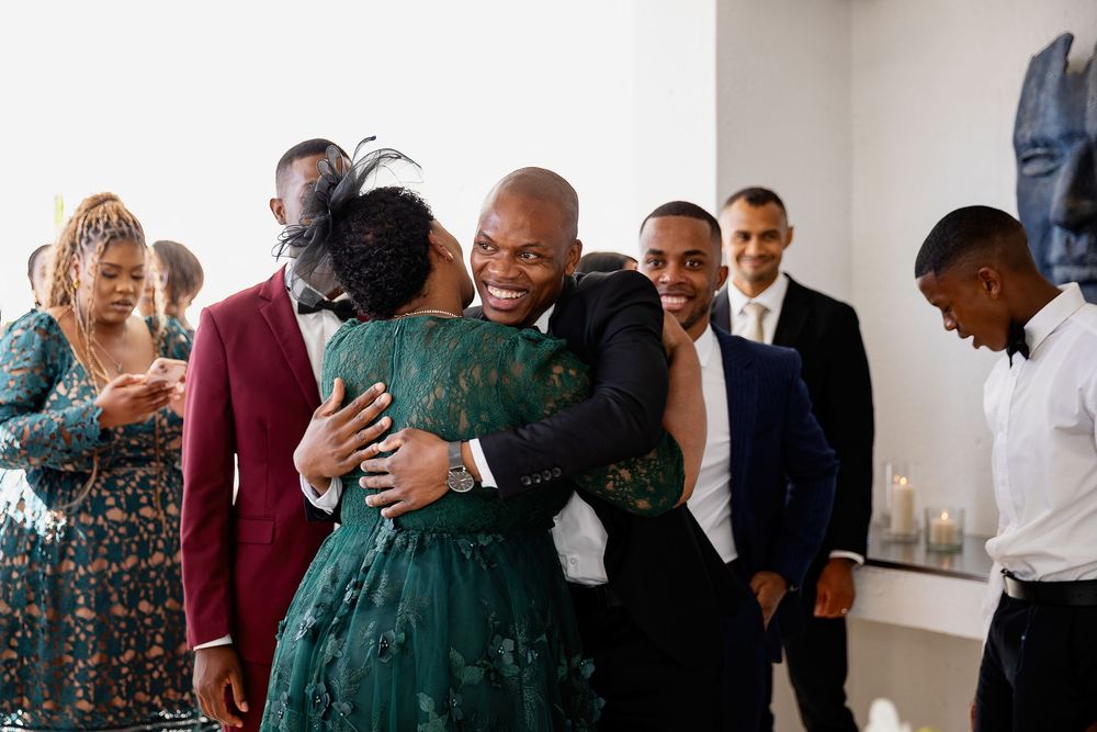 A joyful group of people share warm embraces and laughter during a celebratory gathering in a bright indoor space.