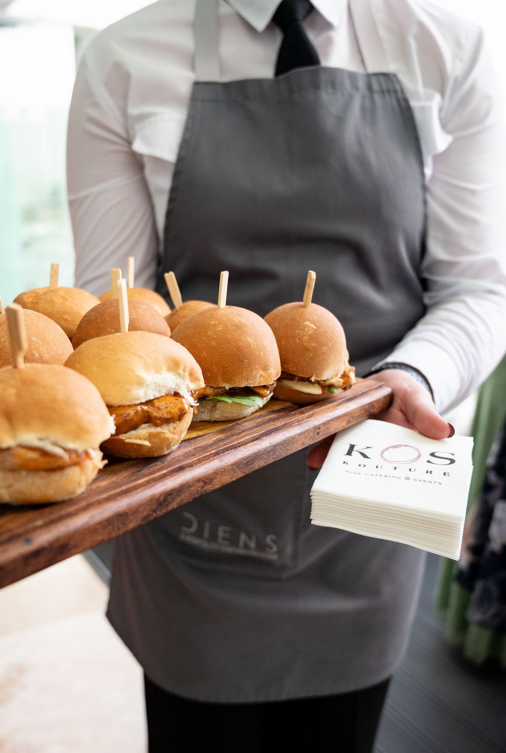 Server holding a wooden board of mini cheeseburger sliders and business cards in a restaurant setting.