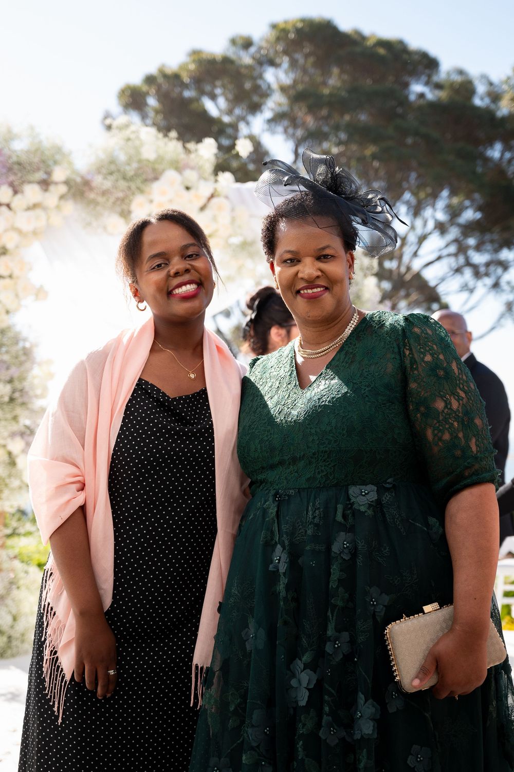 Two women stand together outdoors smiling, one wearing a pink cardigan and polka dot dress, the other in green lace dress.