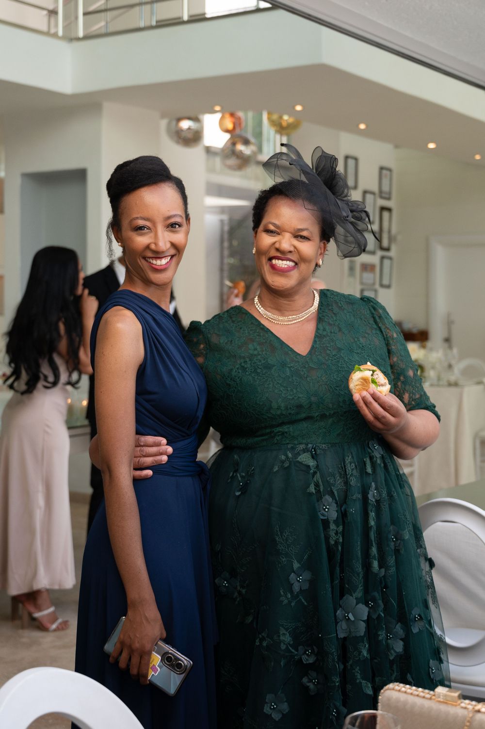 Two elegantly dressed women at a formal event, one in navy blue and one in emerald green lace, stand together smiling.