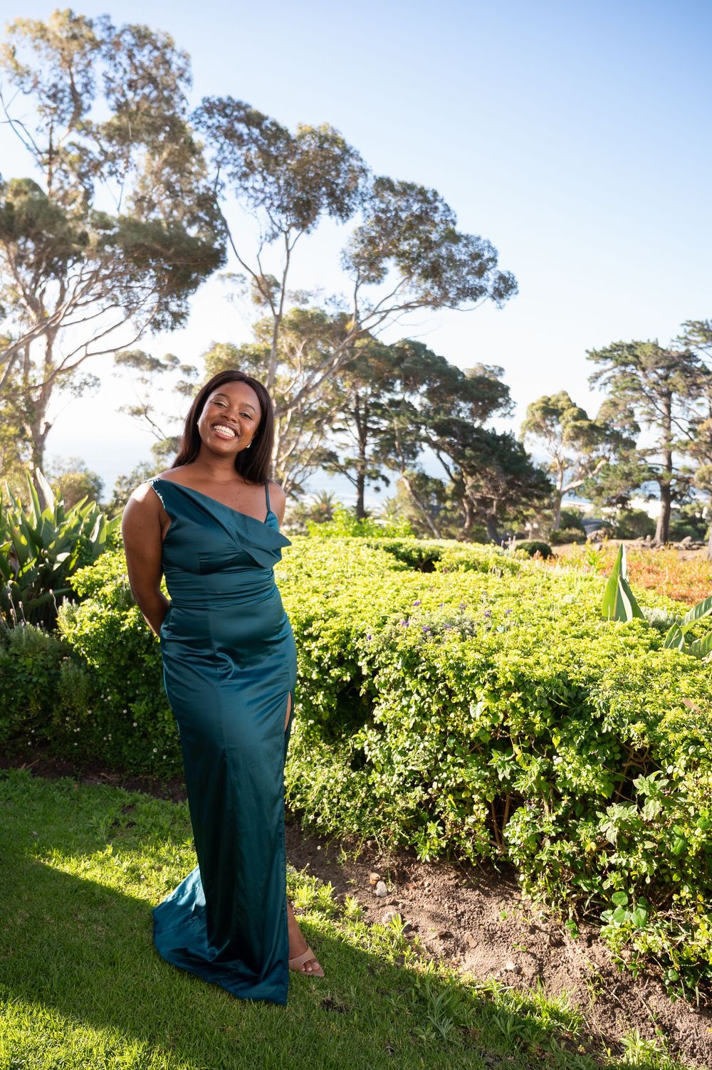 A person in an elegant teal evening gown poses outdoors in a garden setting with tall eucalyptus trees in the background.
