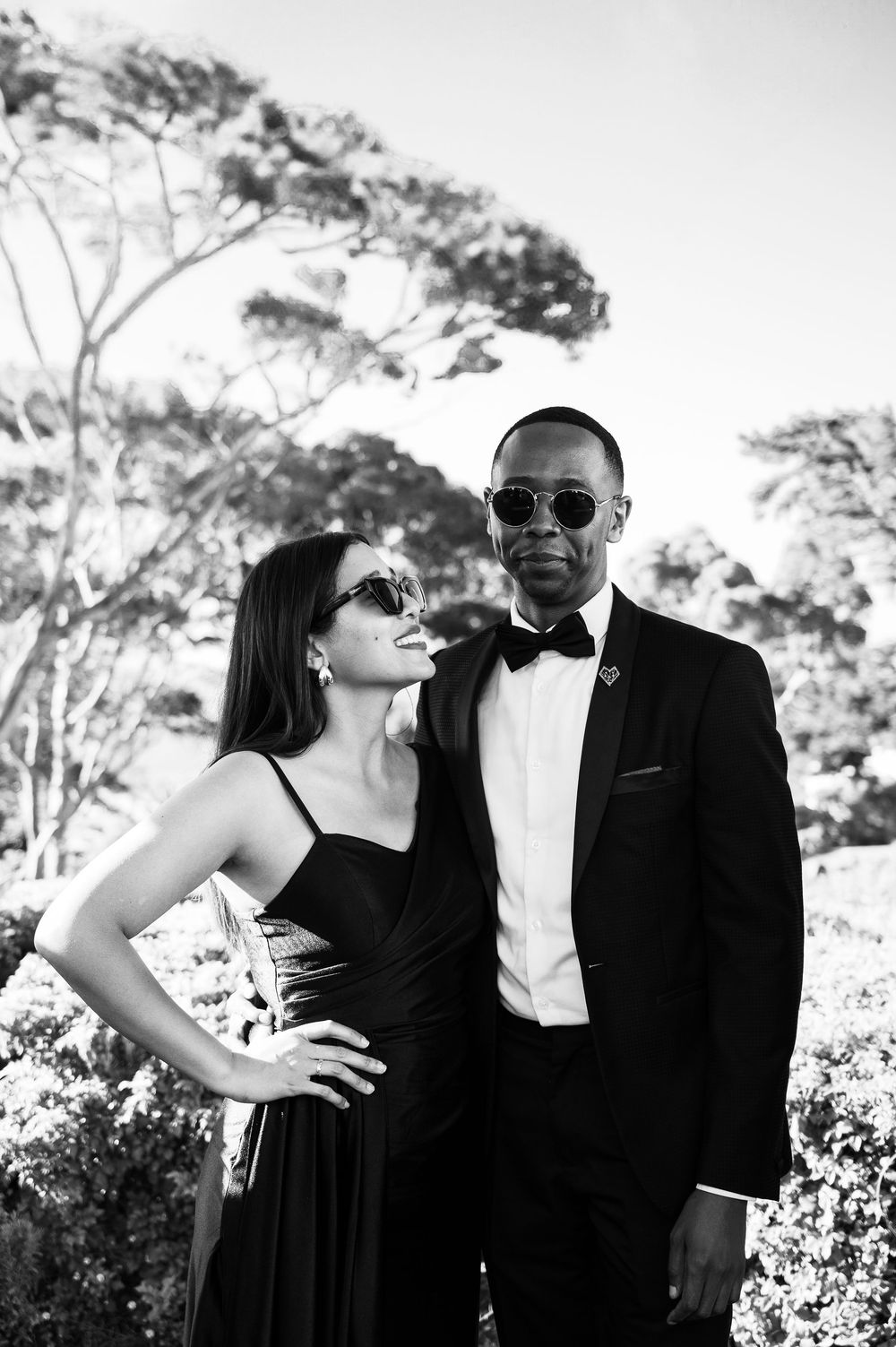 Elegant couple in formalwear with sunglasses pose together outdoors in a black and white photograph with trees.
