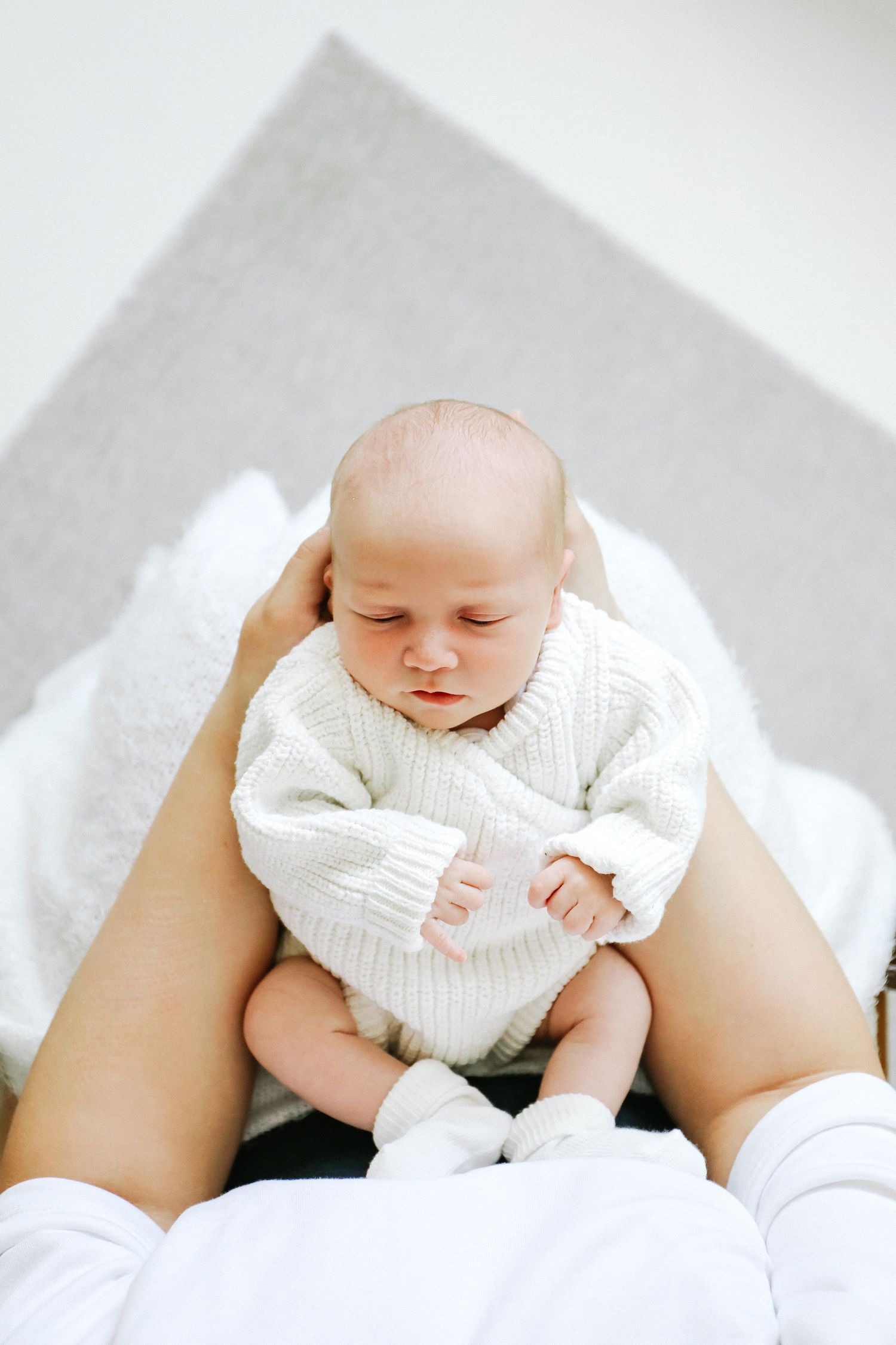 A sleeping infant wearing a cozy white knit rests on mums lap against a light background, during newborn shoot in Nottingham