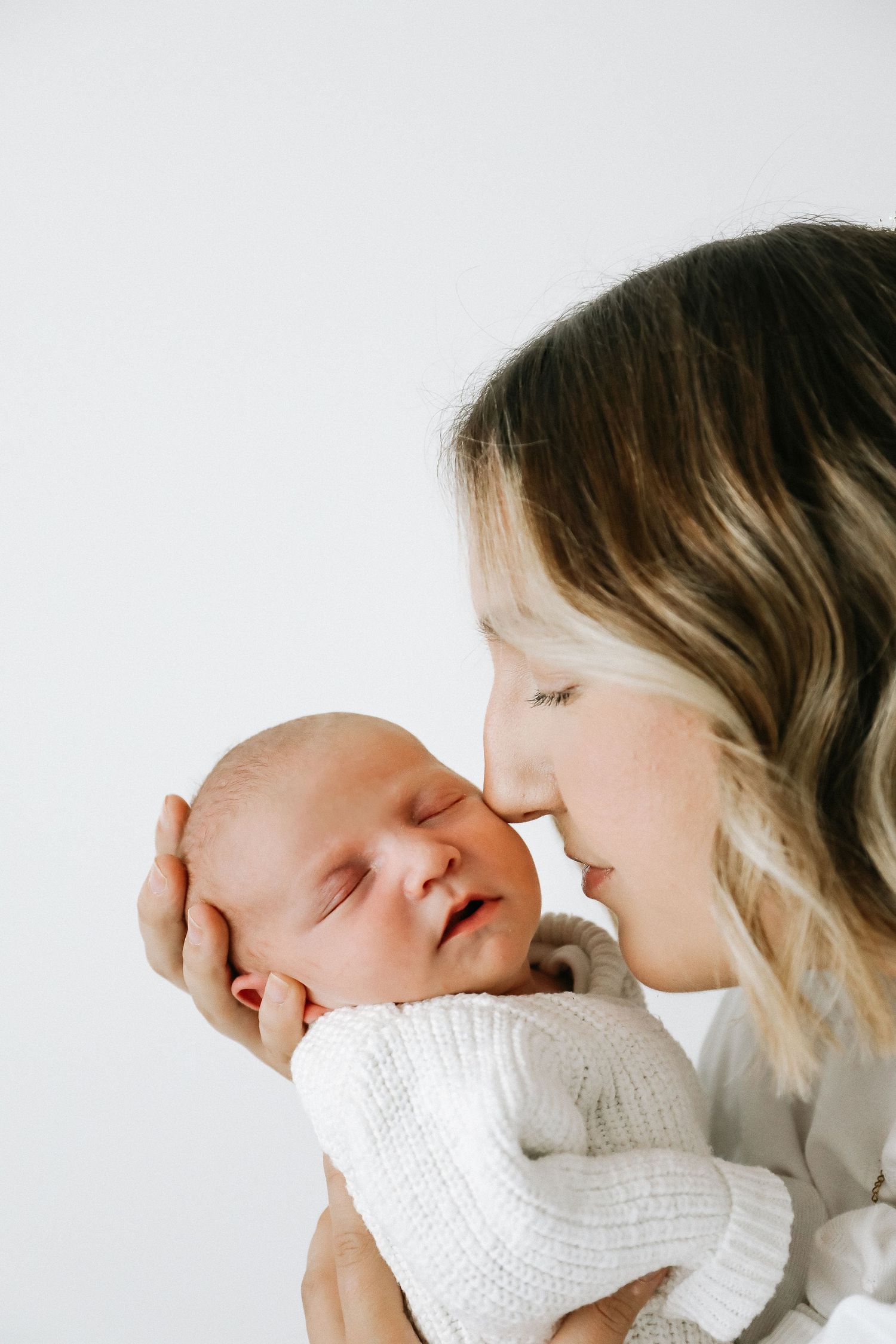 A tender maternal moment shows a mum gently nuzzling a sleeping newborn baby in a white knit sweater, during newborn shoot in Nottingham