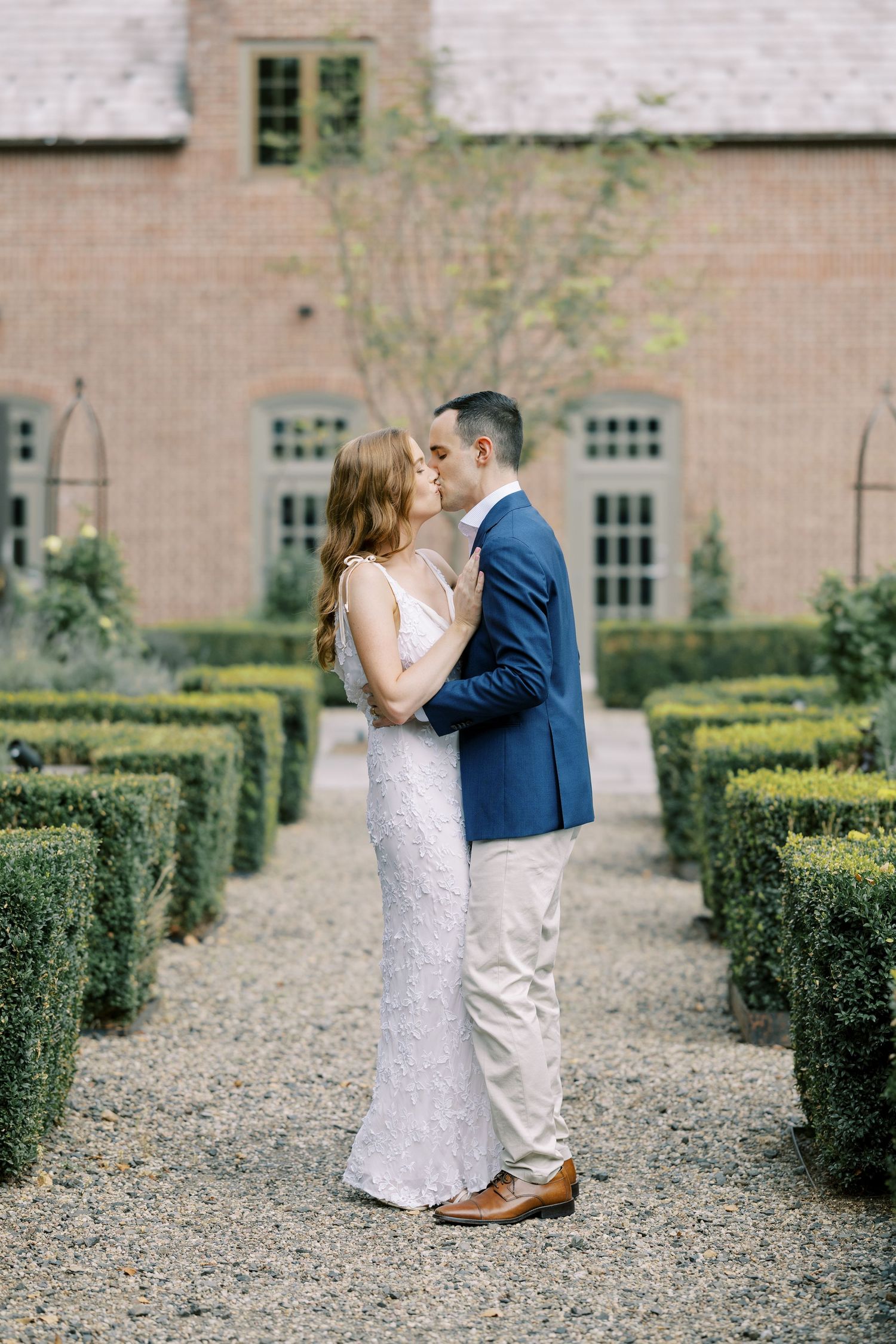 Two people walk together on a gravel path beside a manicured garden and brick building during an outdoor photo session at Ninety Acres.