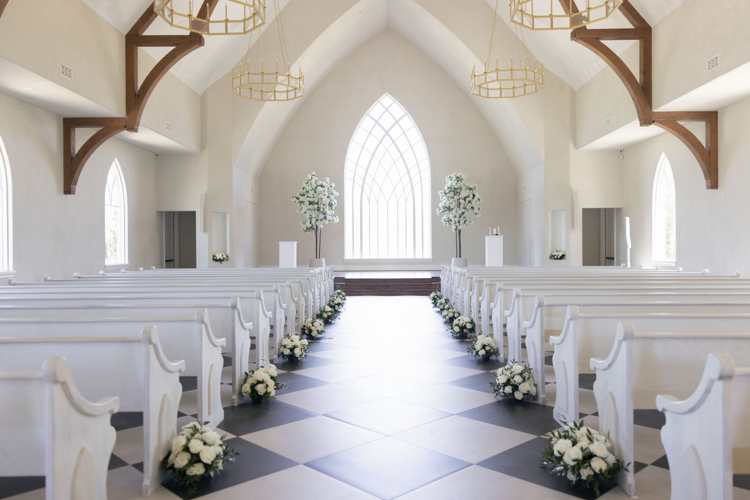 Interior of an elegant white chapel with rows of pews decorated with floral arrangements leading to an arched altar.