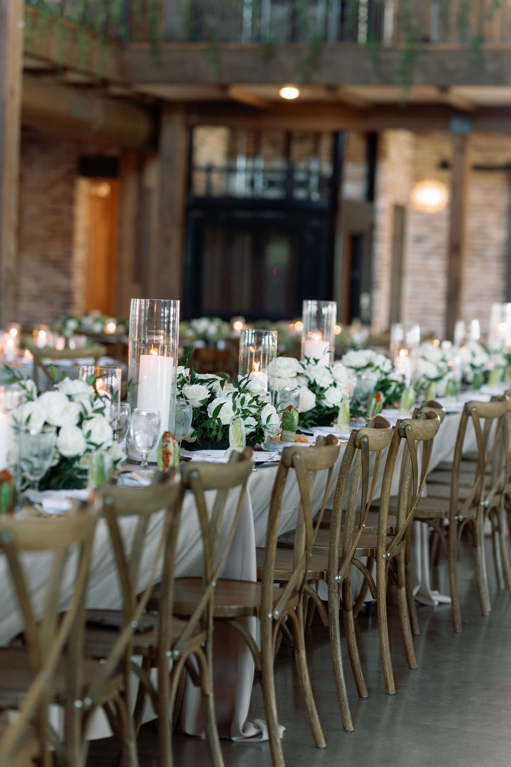 Elegant wedding reception table setting with white floral centerpieces, candles and wooden cross-back chairs in rustic venue.