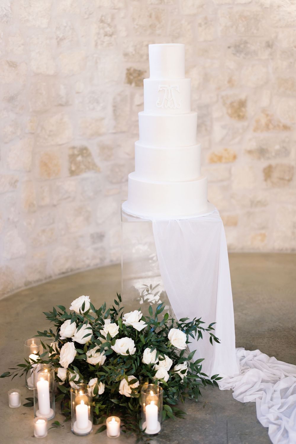 Elegant white tiered wedding cake with greenery and candle decorations against stone wall.
