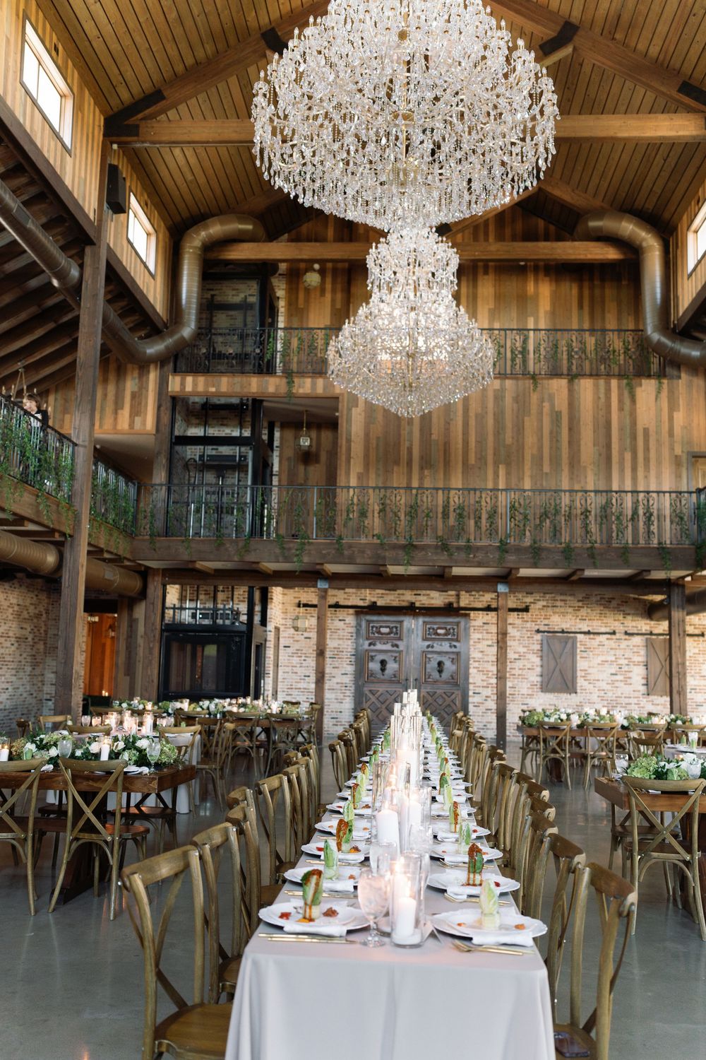 Elegant rustic barn venue with grand crystal chandeliers and long dining tables for a wedding reception.