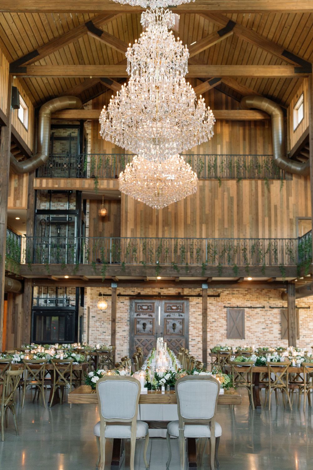 Elegant rustic wedding venue with two tiered crystal chandeliers and wooden barn interior adorned with floral decorations.