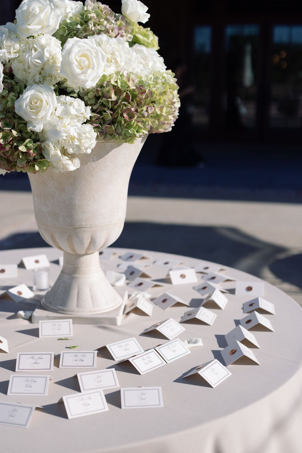 White roses and hydrangeas in stone urn vase with scattered place cards on round table.