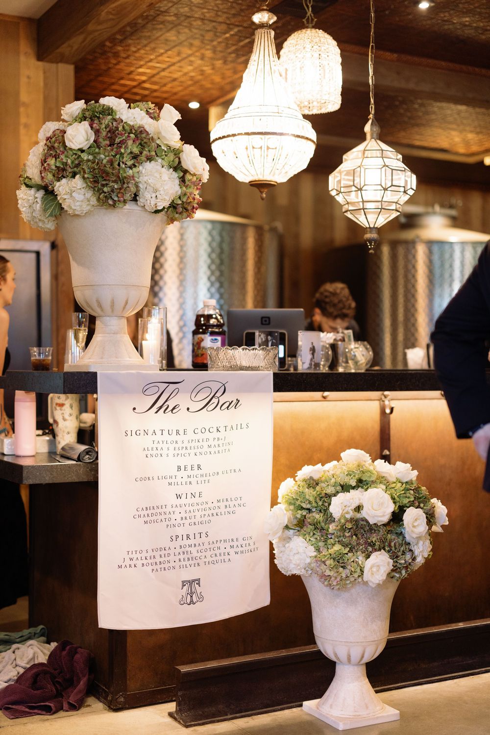 Elegant white floral arrangements in urns and bar menus displayed against dark wood with crystal chandeliers overhead.