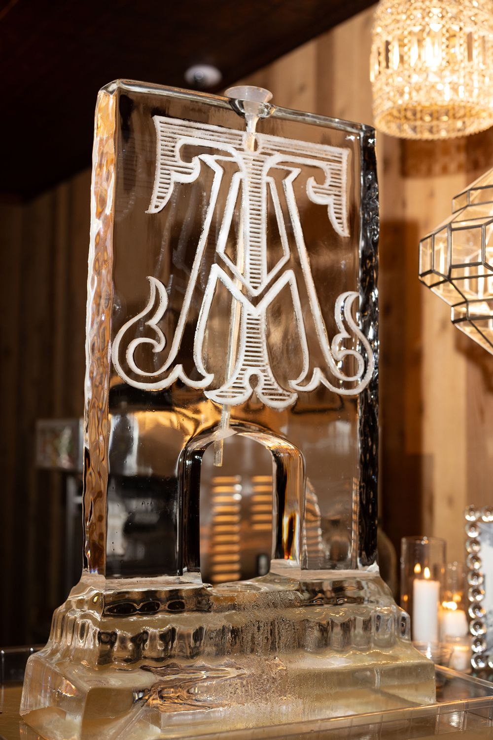An elegant ice sculpture featuring an ornate monogram letter 'A' design illuminated by warm lighting and candles.