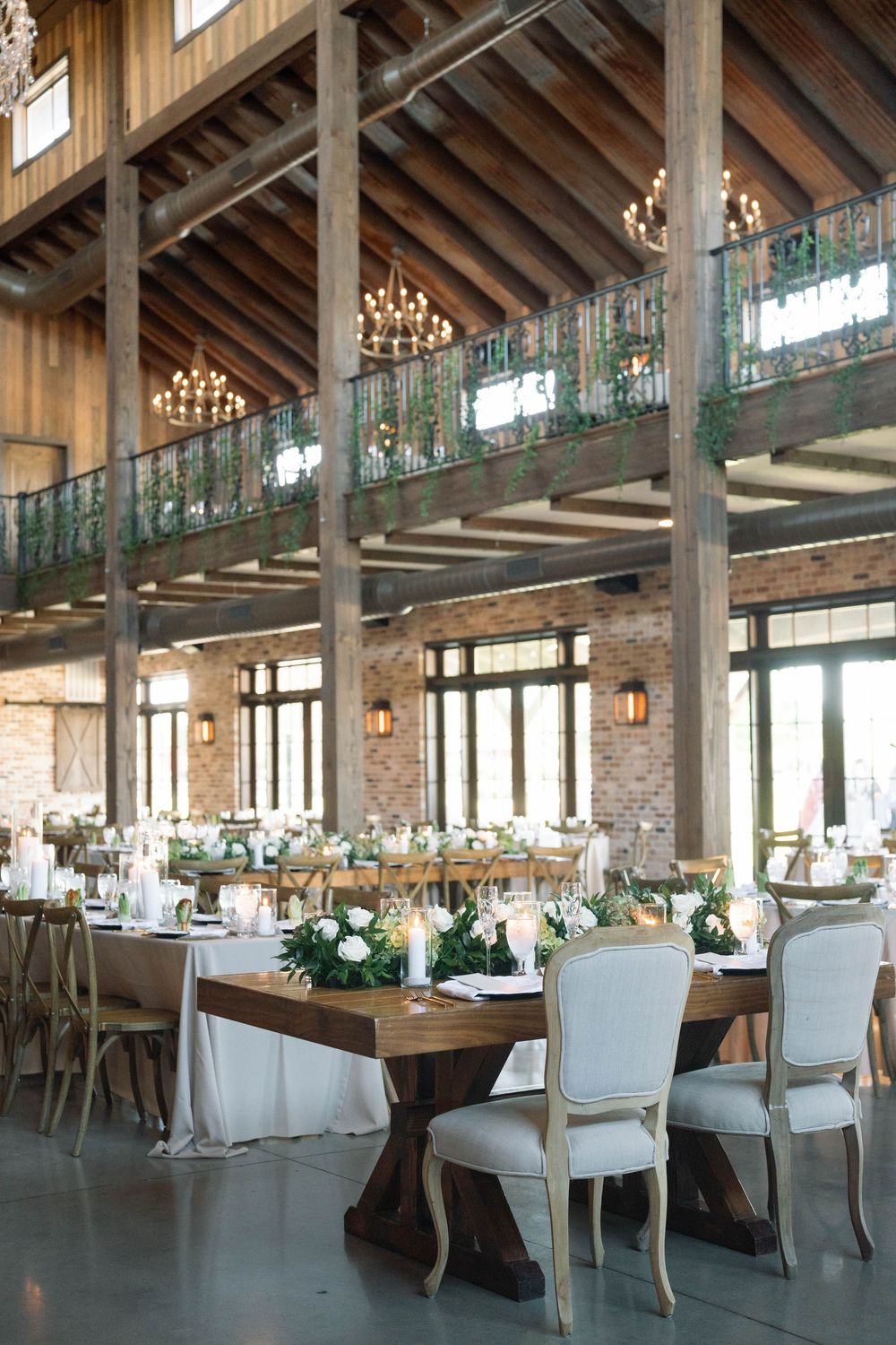 Elegant rustic wedding reception venue with wooden beams, chandeliers, and French country chairs at farmhouse tables.