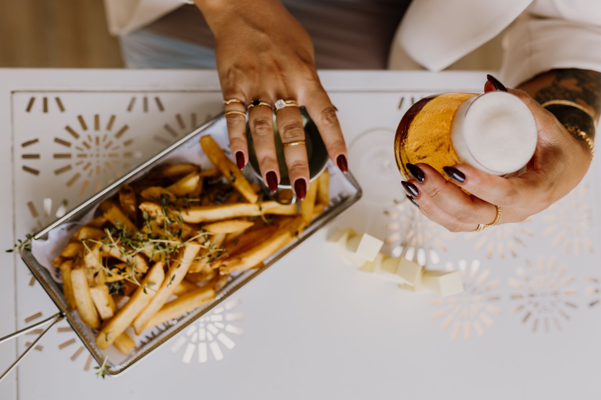 Hands with red nails holding a glass of beer and bowl of French fries.