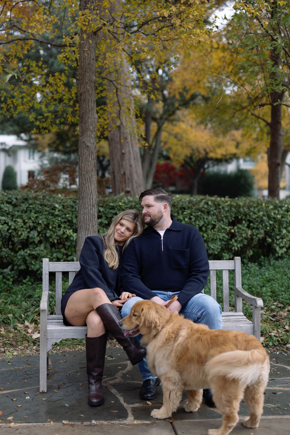 A couple sits on a park bench with their golden retriever on an autumn day surrounded by fall foliage.