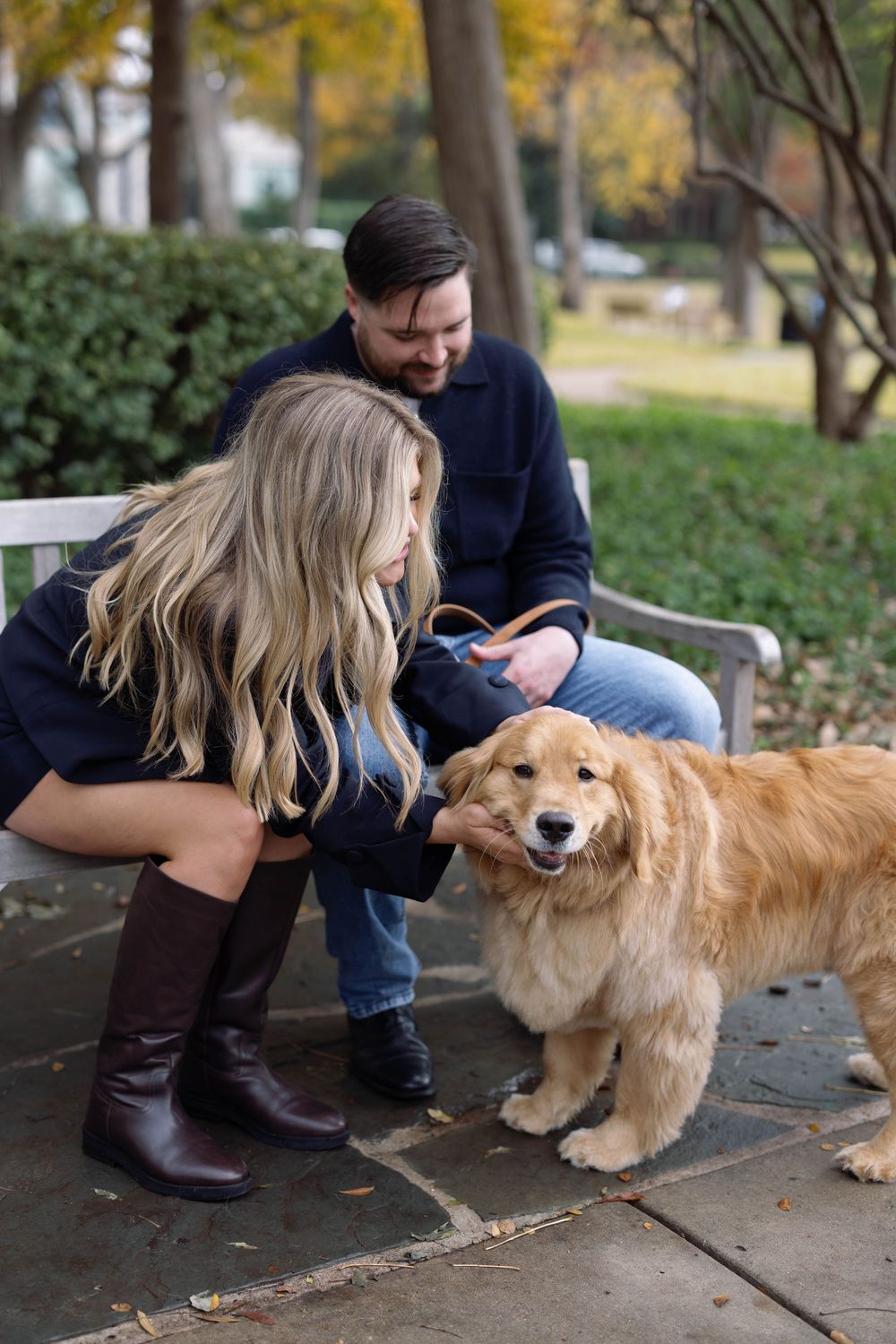 A happy Golden Retriever receives attention from two people sitting on a bench in a park during autumn.
