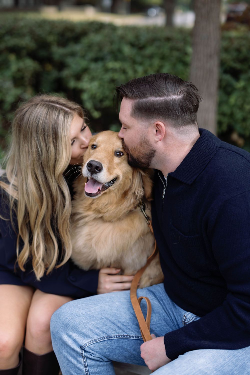 Couple in navy shirts kissing their happy golden retriever mix dog outdoors among green foliage.