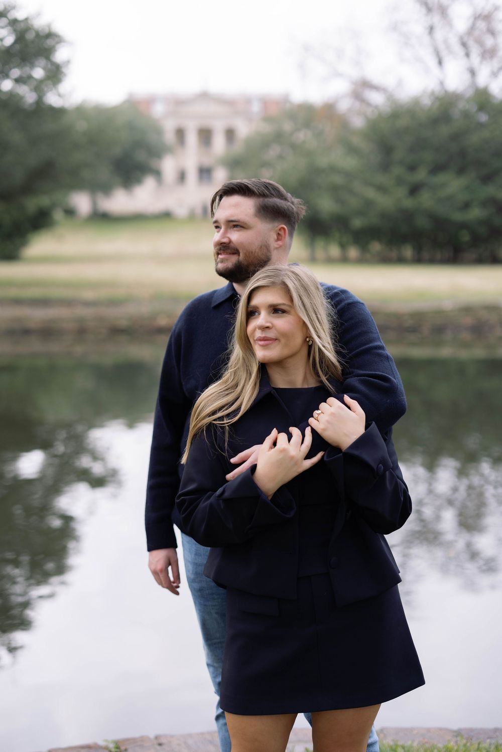 A couple embraces in front of a tranquil pond with a stately building visible in the background on a cloudy day.