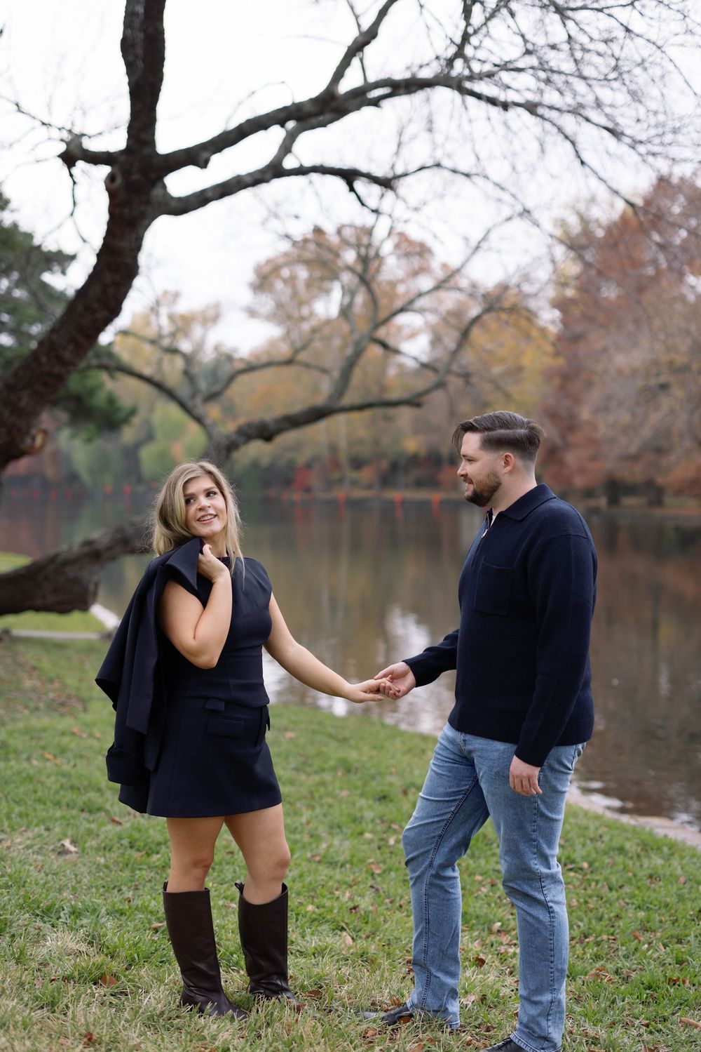 A couple shares a romantic moment by a serene lake on an autumn day with fallen leaves and bare tree branches.