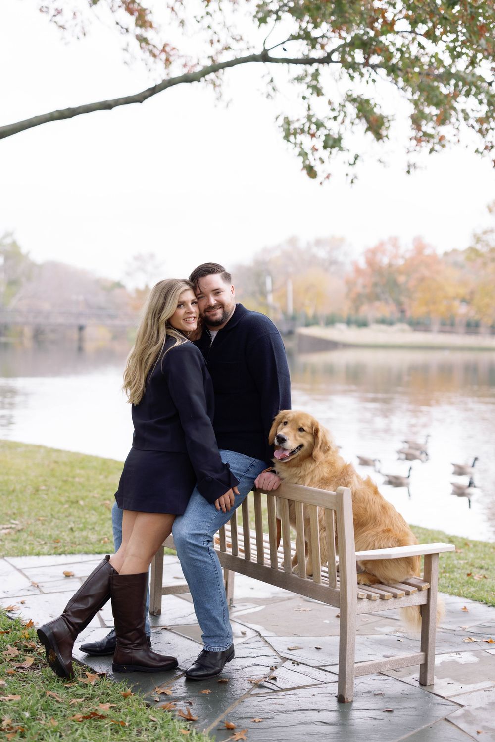 A couple poses with their golden retriever on a wooden bench by a serene lake in fall.