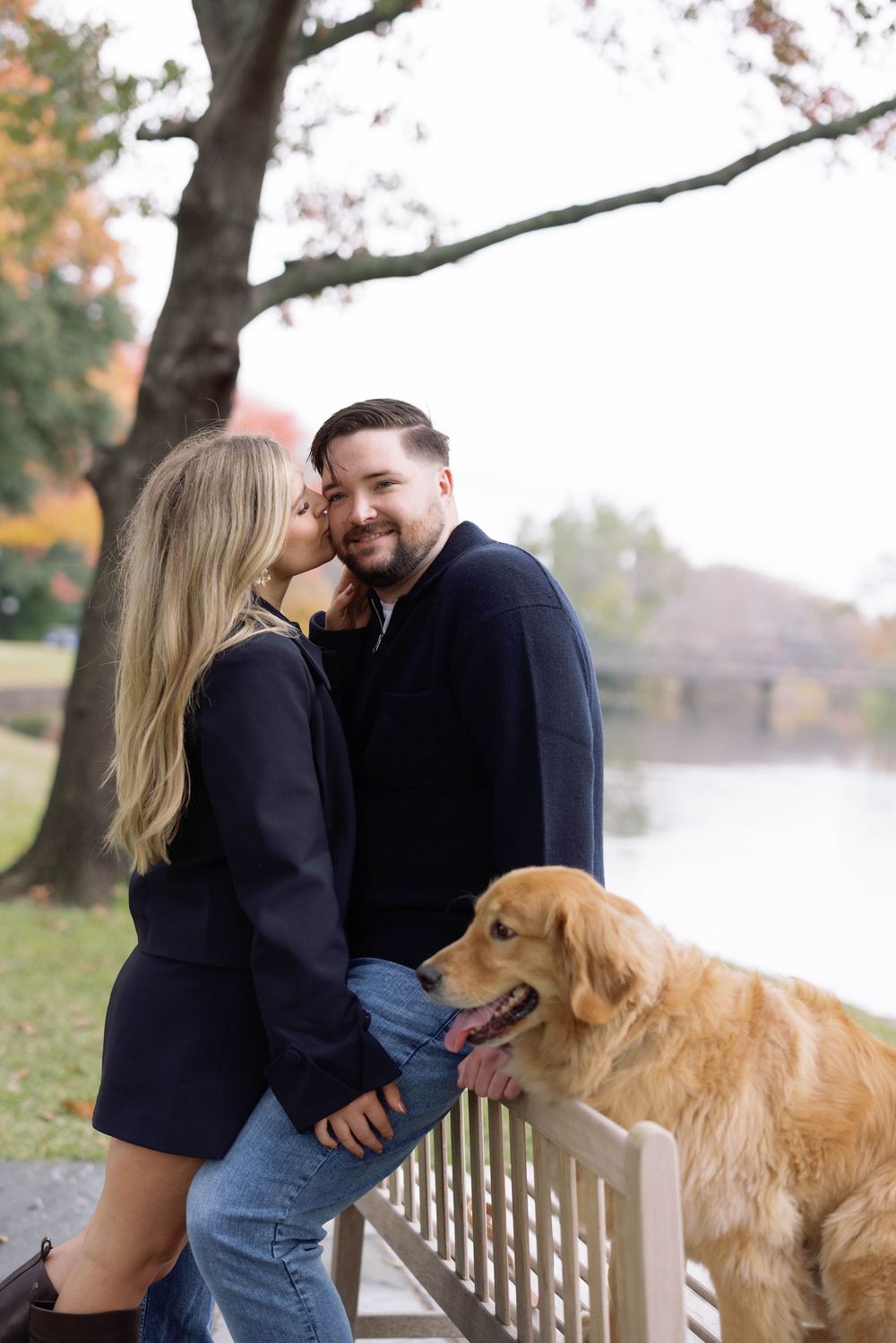 A couple shares a tender moment with their golden retriever by a lakeside during fall.