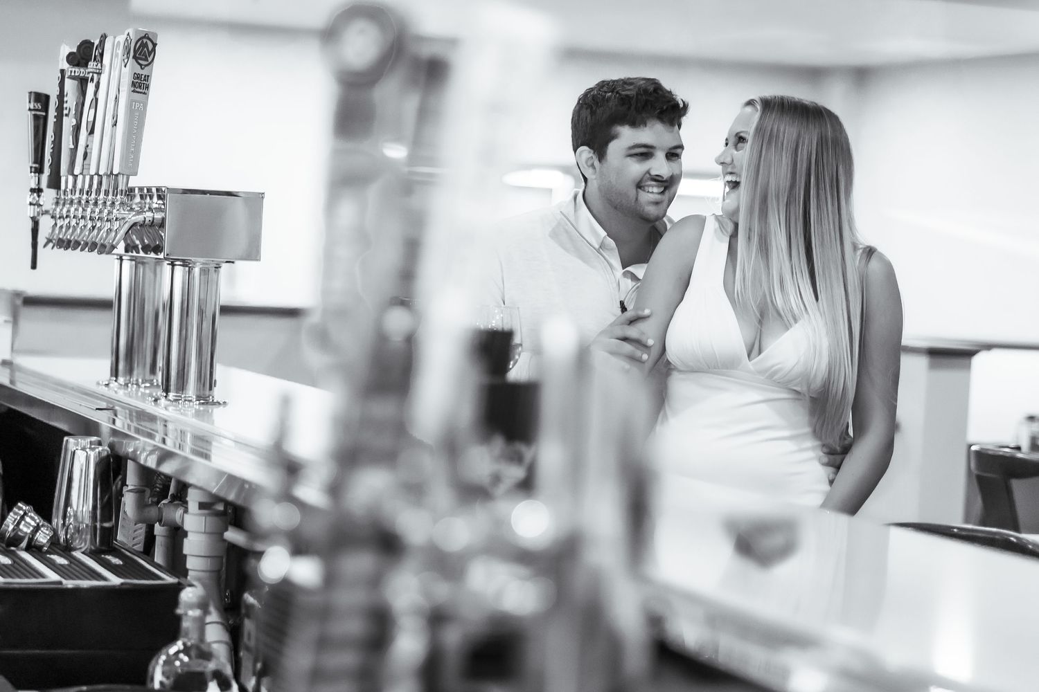 A romantic couple laughs together while standing at a bar counter with draft beer taps in the foreground.