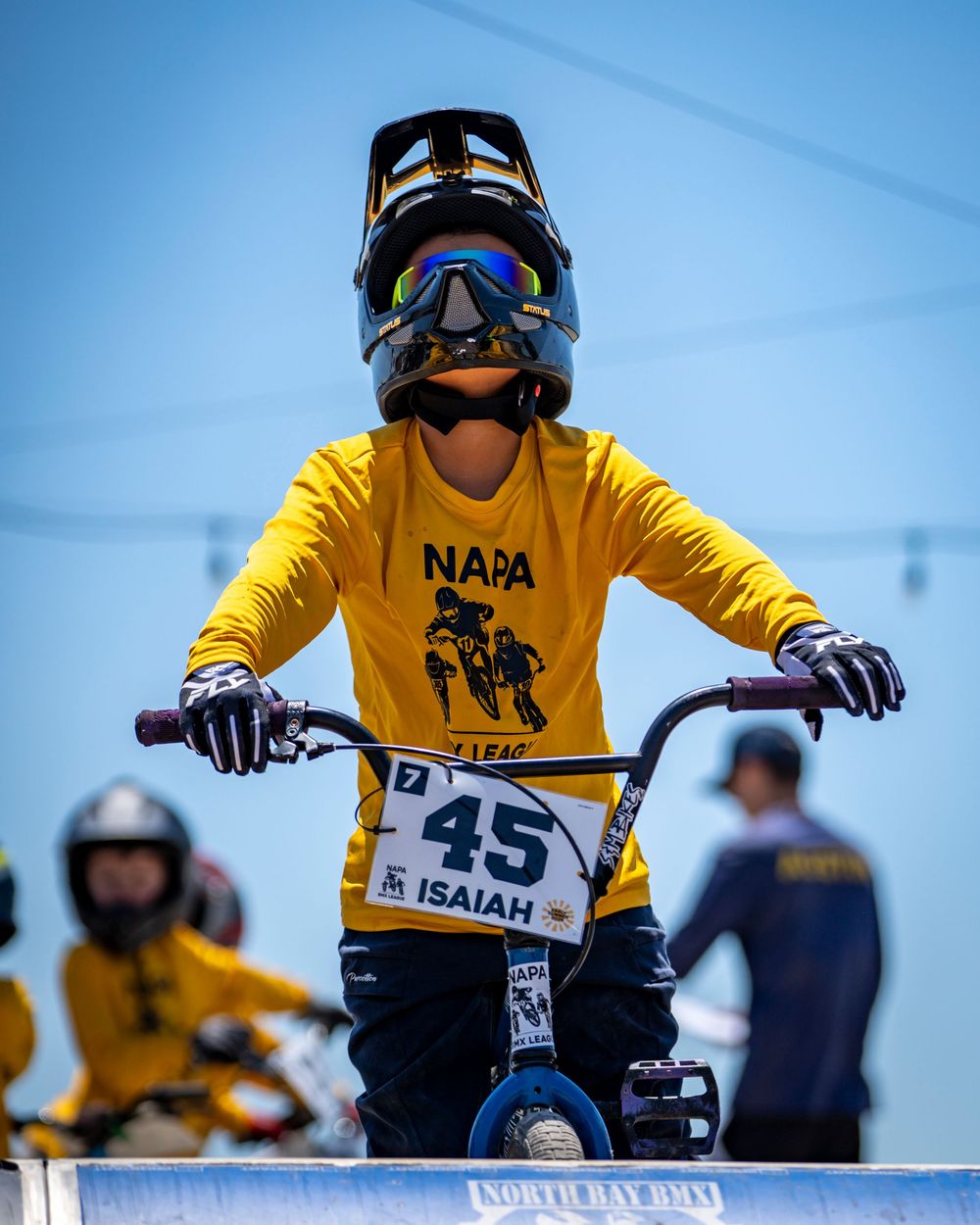 BMX rider in yellow NAPA jersey and protective gear racing on outdoor track.