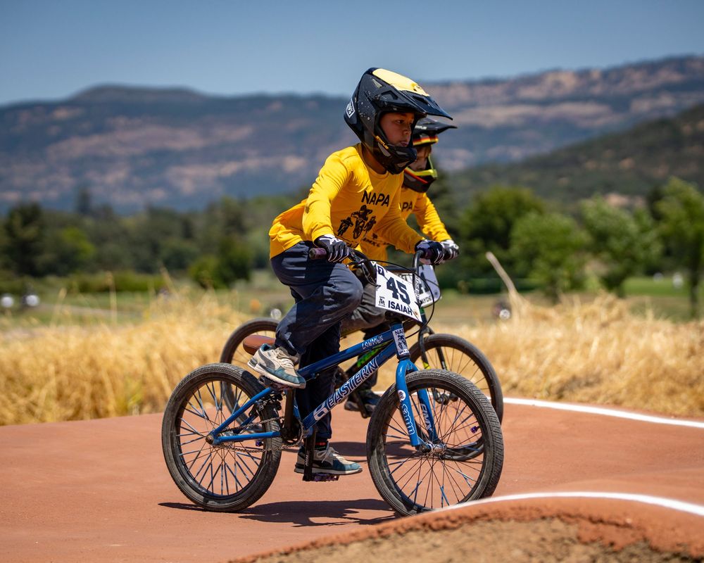 BMX racer in yellow jersey rides on dirt track with mountain landscape in background.