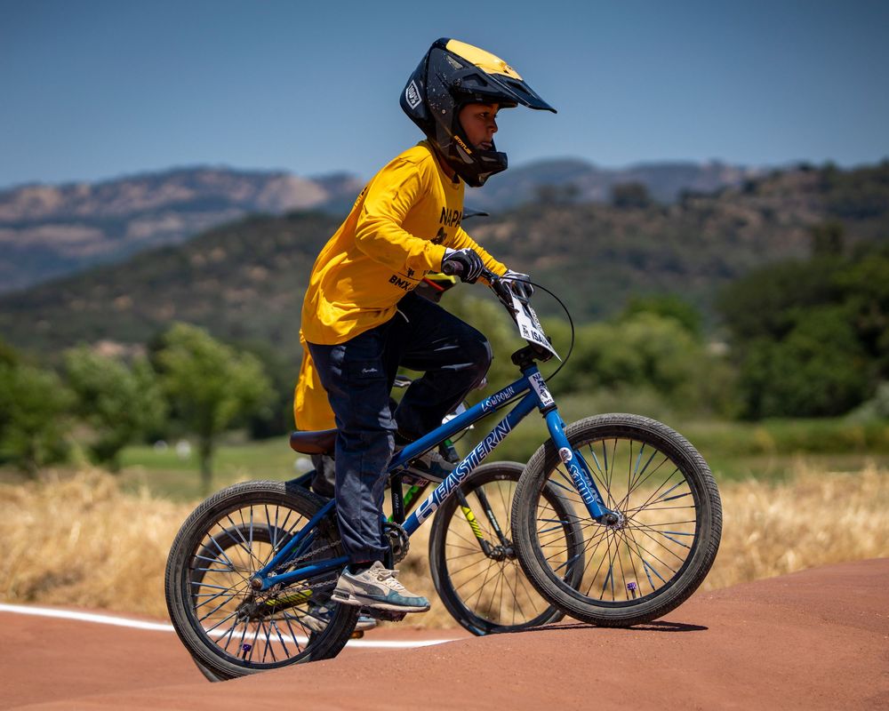 BMX racer in bright yellow jersey navigates dirt track on blue bicycle.
