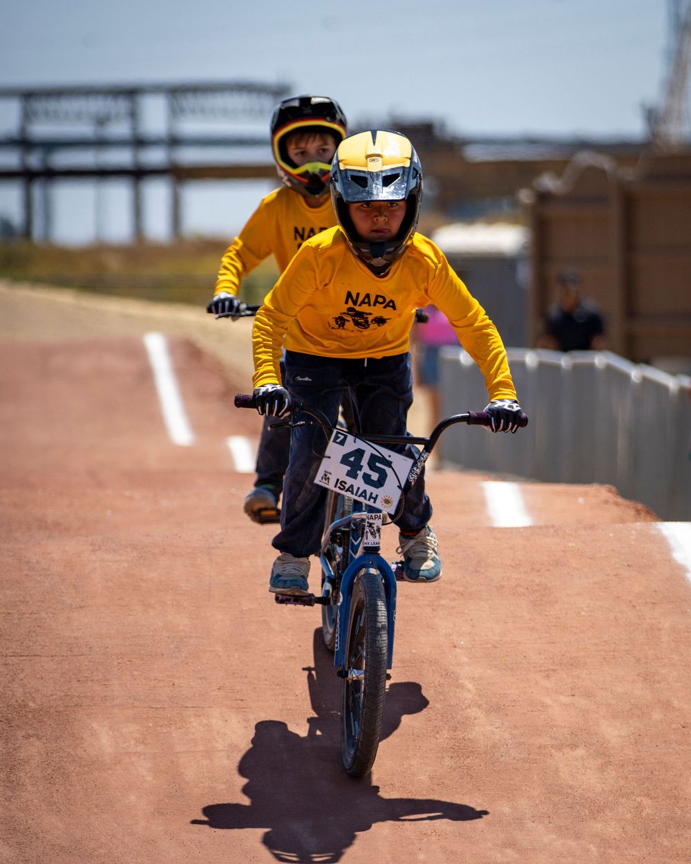 Two BMX riders in yellow NAPA jerseys racing on outdoor track.
